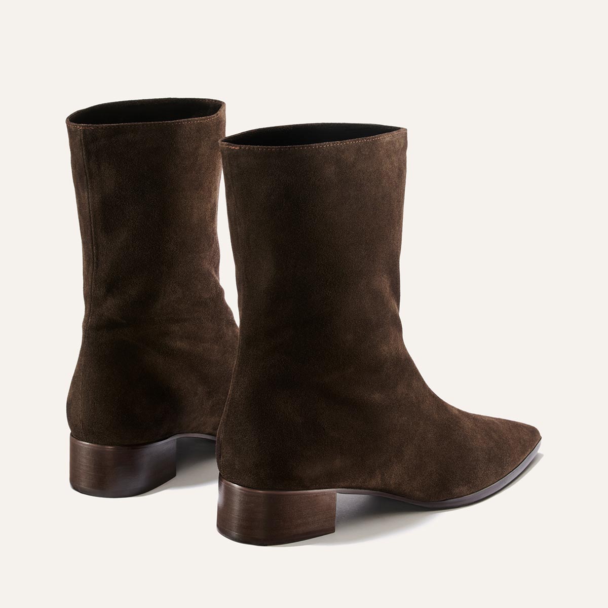 A pair of Margaux’s The Hayes Boot in dark chocolate suede, featuring pointed toes and low, wide wooden heels, is displayed against a light background and shown from the back and side angles, exuding a subtle bohemian flair.