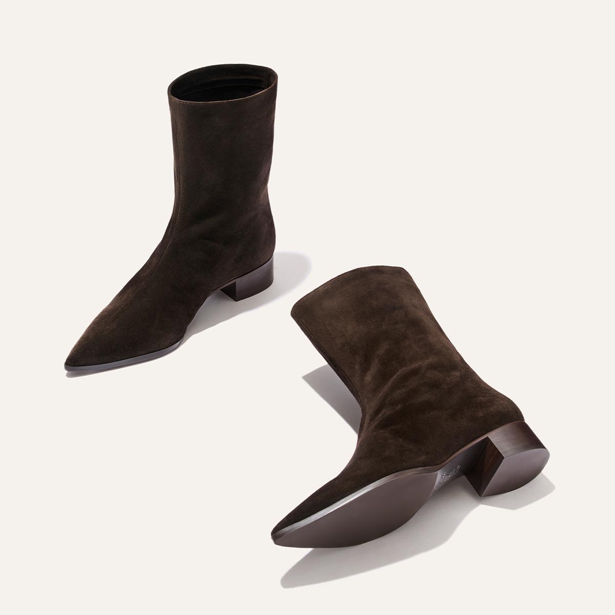 The Hayes Boot by Margaux in Dark Chocolate Suede is a pair of soft, mid-calf boots with low block heels and pointed toes, offering a subtle bohemian flair, displayed on a light background.