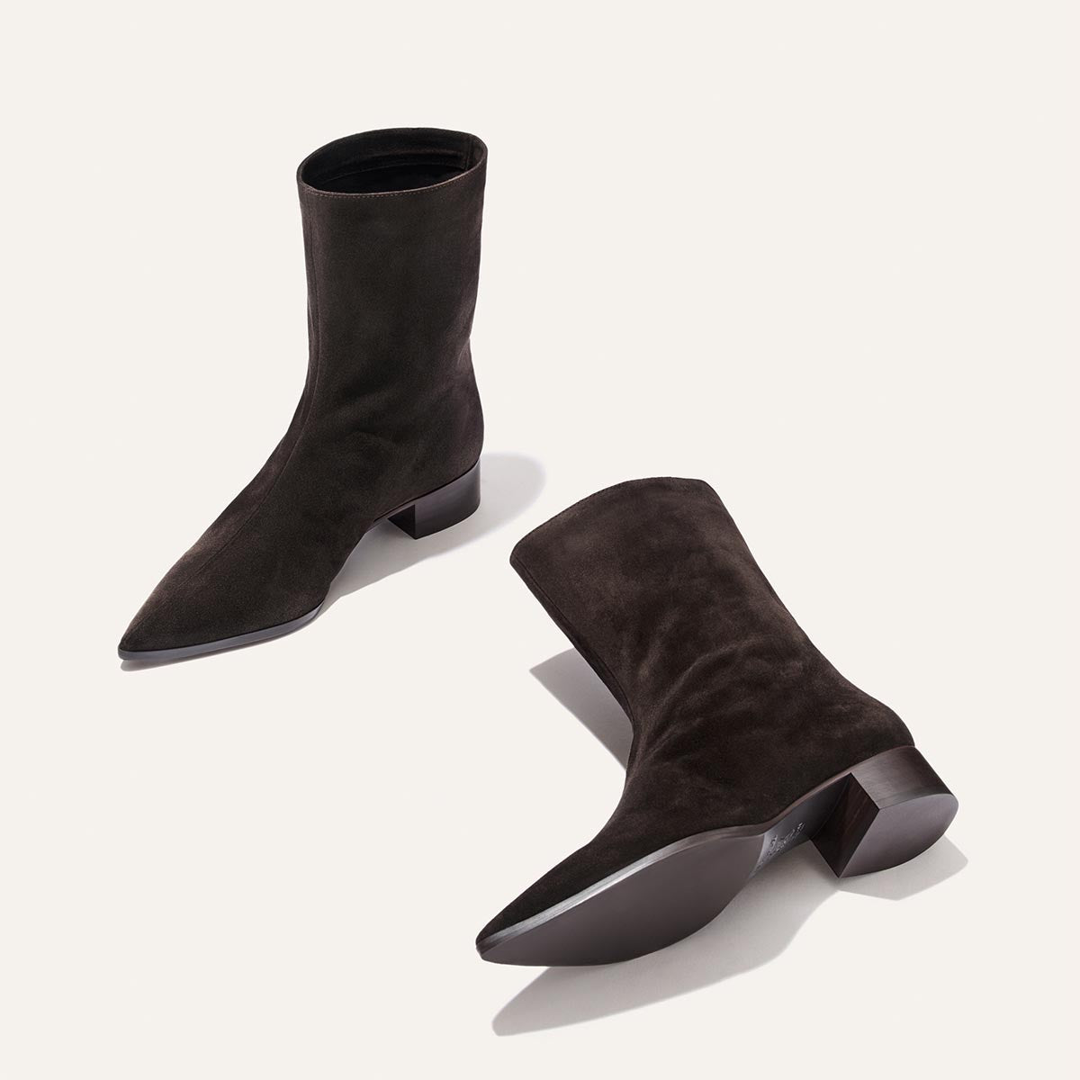 The Margaux Hayes Boot in Dark Chocolate Suede features pointed toes and low block heels; one boot stands upright while the other lies on its side, both displayed against a light background.