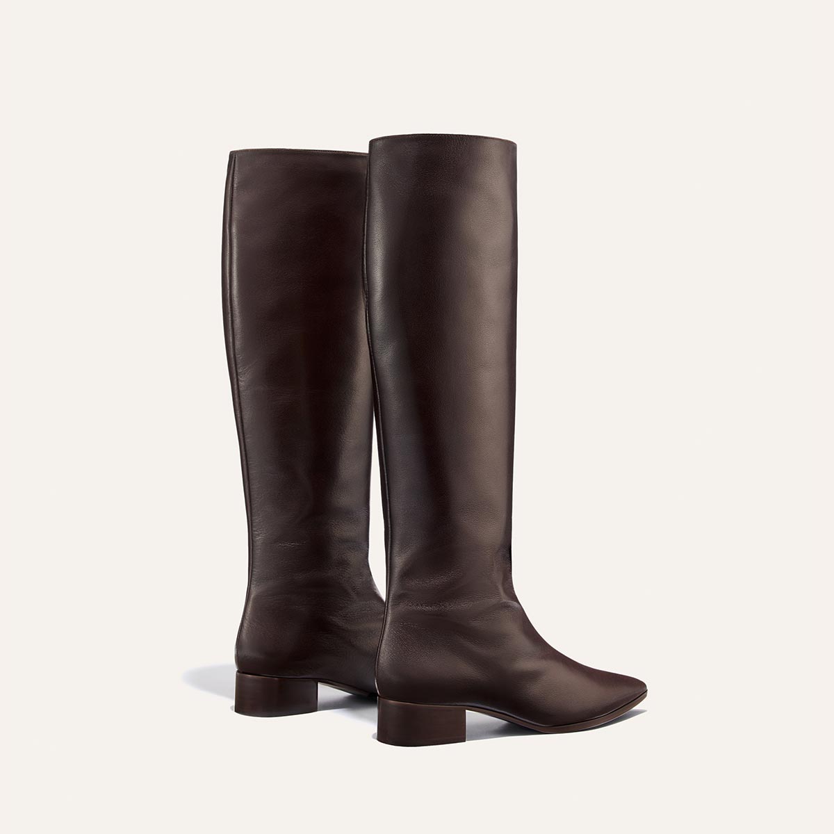 The Hayes Knee-High Boot in Dark Chocolate Calf by Margaux is displayed against a light background. This pair of handmade leather boots features a tall silhouette, low block heels, and a flexible sole for added comfort. One comfortable boot faces forward while the other is angled slightly to the side.