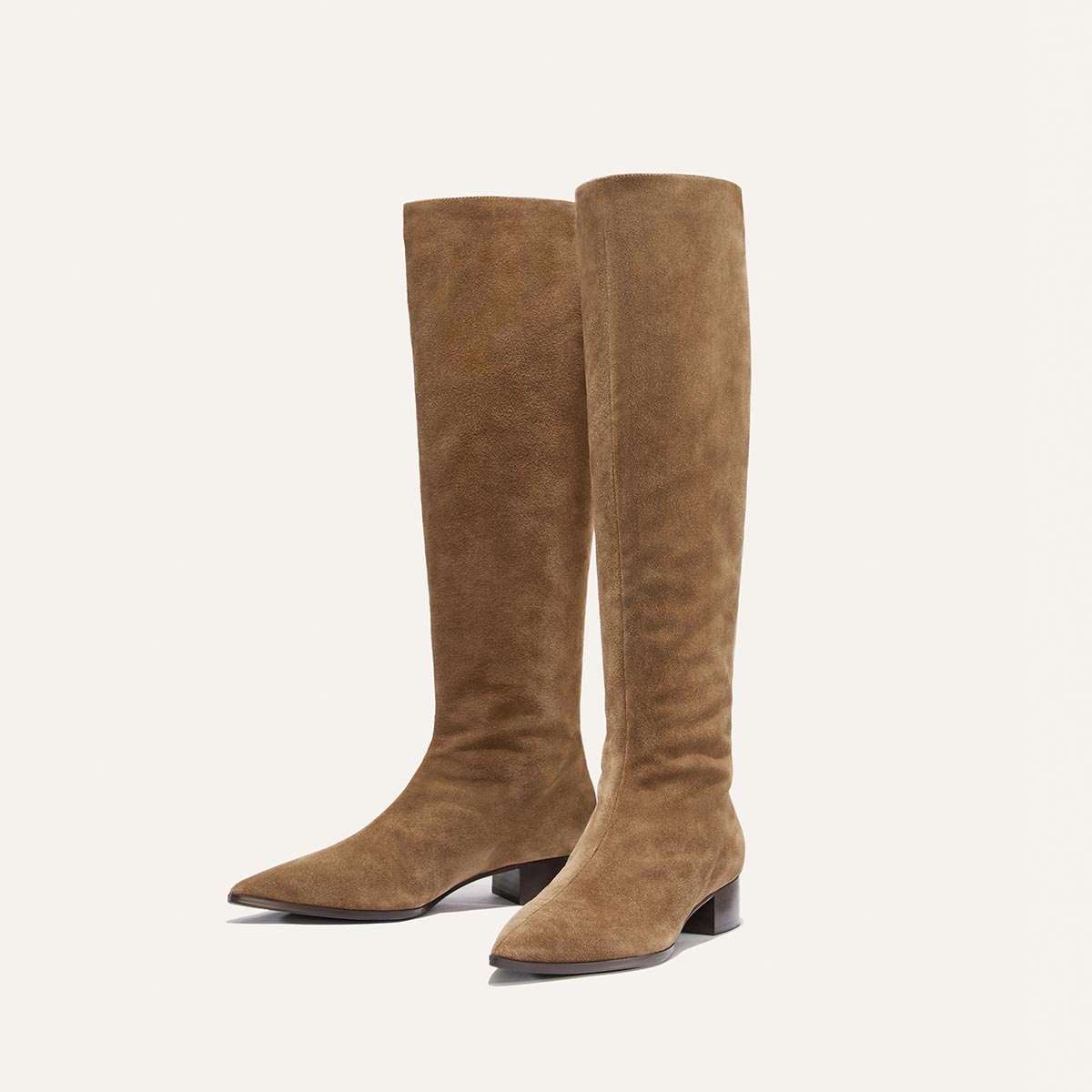 The Hayes Knee-High Boot in Driftwood Suede by Margaux is displayed upright against a plain, light background. These handmade boots feature a tan suede finish, low block heels, and pointed toes. Designed for all-day comfort, they are crafted with leather and have a flexible sole for easy wear.