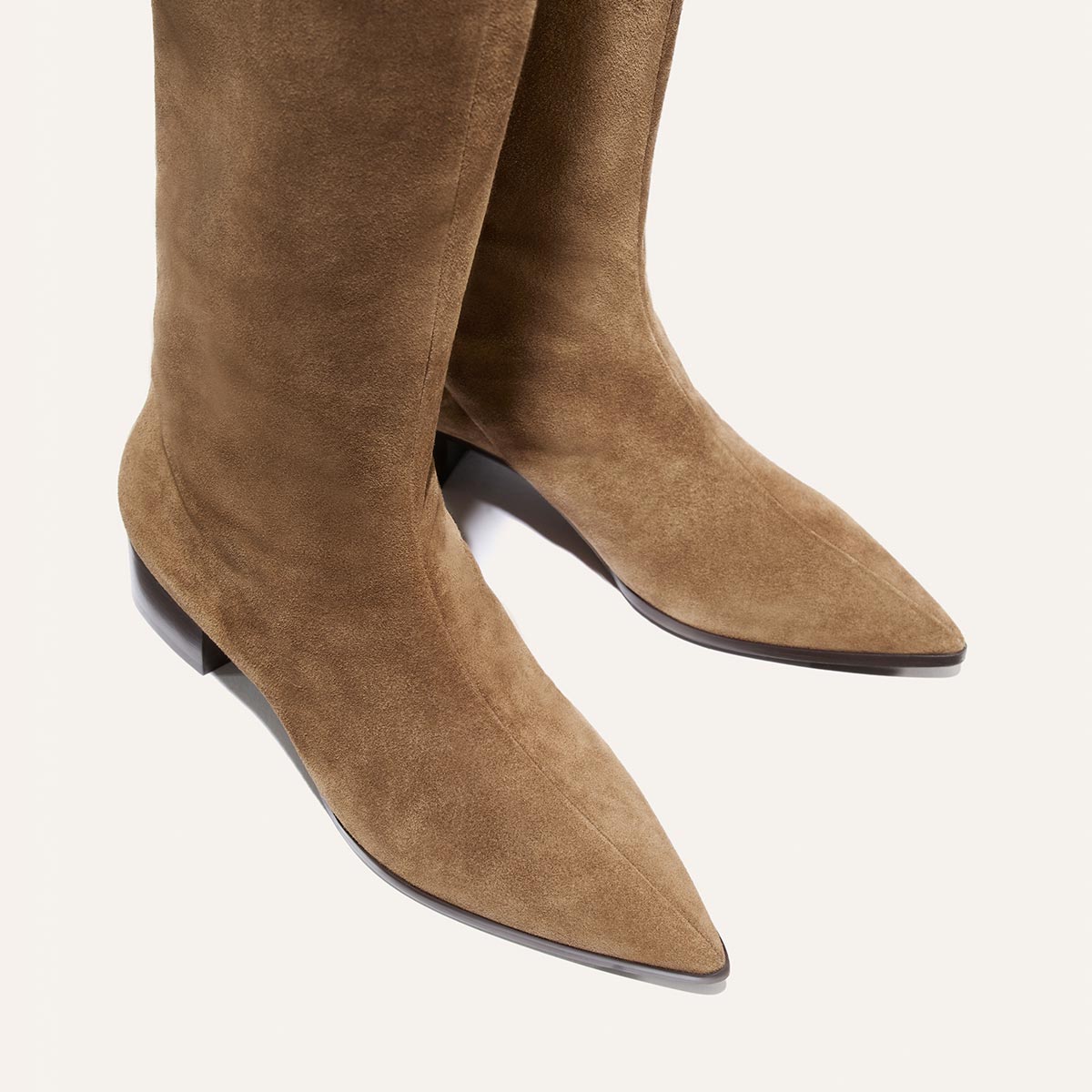 Close-up of The Hayes Knee-High Boot in Driftwood Suede by Margaux—a pair of tan suede boots with pointed toes, low heels, and a flexible sole for added comfort—shown against a plain light background.