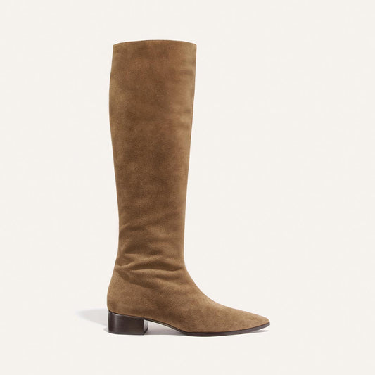 Introducing The Hayes Knee-High Boot - Driftwood Suede by Margaux: a tall, tan suede knee-high boot featuring a low, dark brown block heel, slightly pointed toe, and flexible sole for enhanced comfort. This handmade boot is elegantly displayed against a clean light background.