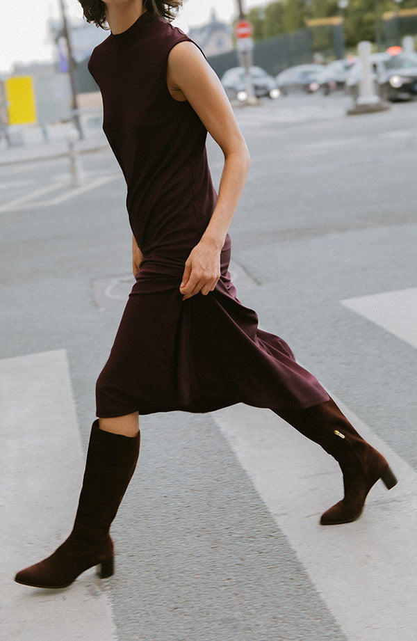 A person in a sleeveless, dark purple dress and matching knee-high boots walks briskly across a city street, holding the hem of the dress. The background shows an urban setting with blurred buildings and traffic signs.