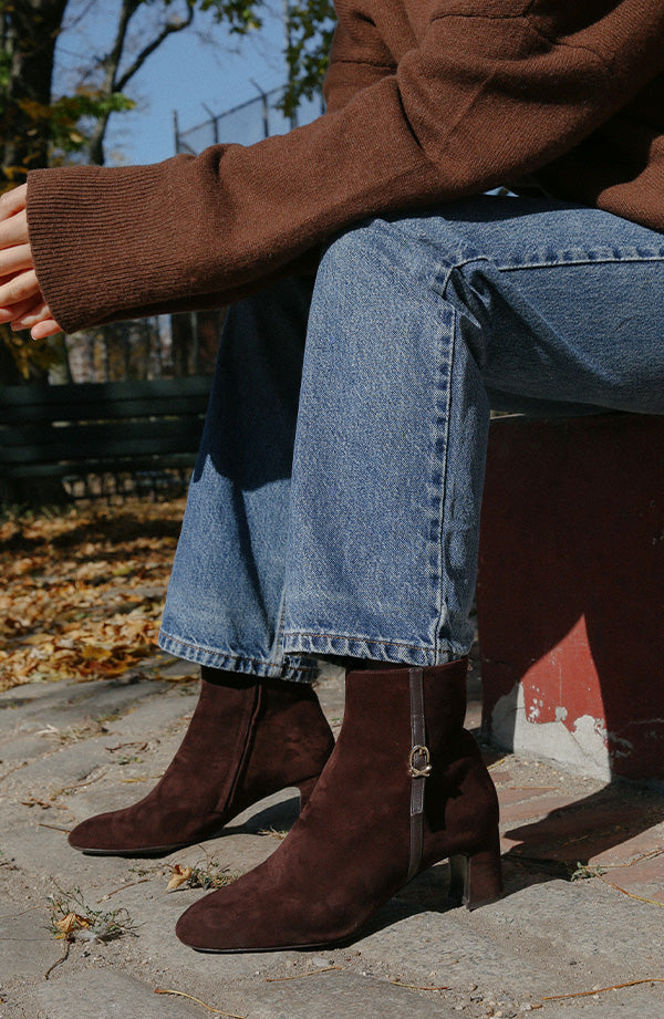 The Bowery Boot in rich chocolate suede, an ankle boot featuring an elegant breasted heel and gold-tone buckle hardware.