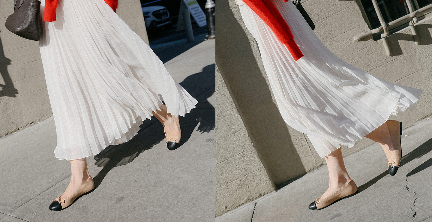 A person wearing a flowing white pleated skirt and beige flats with black toe caps is walking on a sunny sidewalk; their upper body is out of frame.
