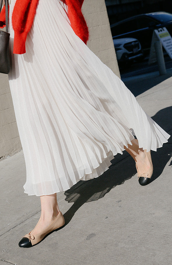 A person wearing a flowing white pleated skirt, black-toed beige ballet flats, and a red sweater draped over one arm walks on a sunny sidewalk.