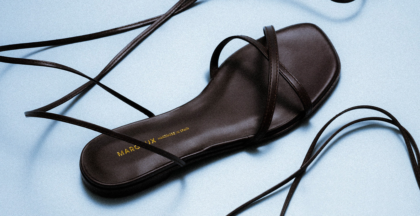 A single black strappy sandal with long laces lies on a light blue surface. The sandal has a flat sole and MARA MIX with additional text in gold inside the footbed.
