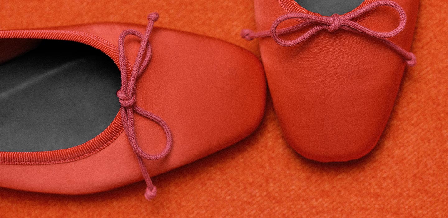 Close-up of a pair of red ballet flats with matching bows on a textured red surface, showing part of one shoe’s insole and the tops of both shoes.