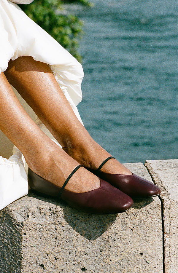 Brown shoes with thin straps worn by a person sitting by a body of water.