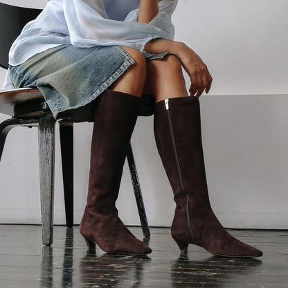 A person sits on a wooden chair, wearing a light blue blouse, a knee-length denim skirt, and the Margaux "The Imogen - Espresso Suede" knee-high boots, set against a plain white wall and dark wooden floor.