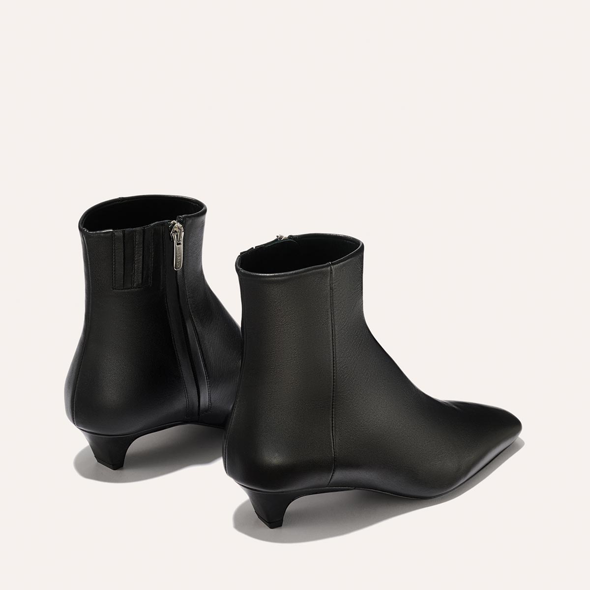 A pair of Alex Mill x Margaux: The Inga - Black Calf ankle boots by margaux-development, crafted in black leather with low kitten heels and side zippers; one boot displays the zipper, the other shows a sleek NYC-inspired silhouette.