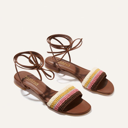 The Isla Wrap Sandal by Margaux features flat brown sandals with ankle wrap laces and a front strap in multicolor crochet—cream, yellow, pink, and brown—with a gold Margaux logo on the insole.