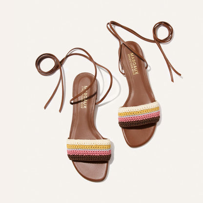 The Isla Wrap Sandal by Margaux features brown flats with long ankle ties and multicolored crocheted straps in white, yellow, pink, and burgundy, shown against a white background.