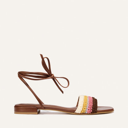 The Isla Wrap Sandal by Margaux features flat brown sandals with ankle wrap laces and a front strap in multicolor crochet—cream, yellow, pink, and brown—with a gold Margaux logo on the insole.