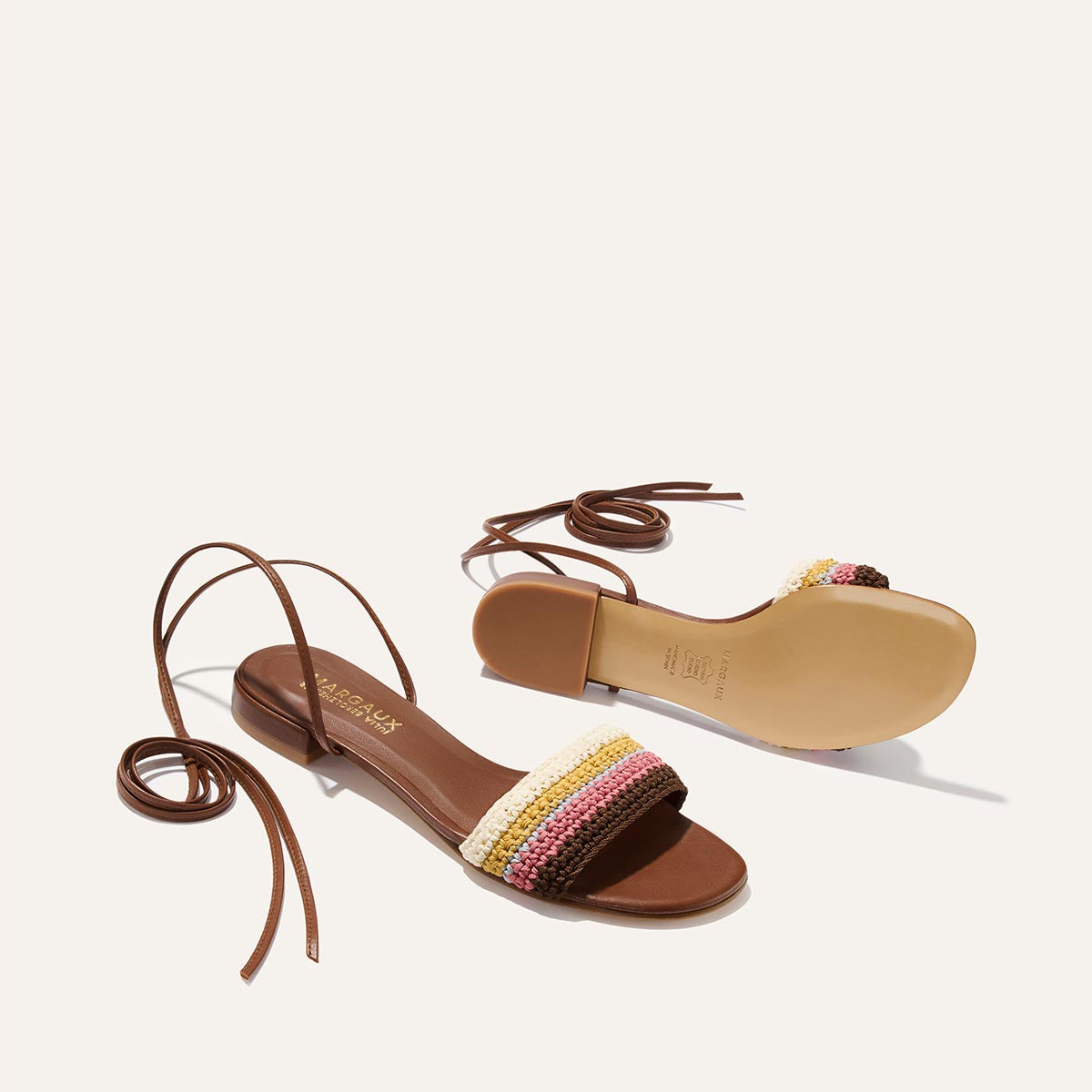 The Isla Wrap Sandal by Margaux in Cognac features flat soles, long ankle straps, and multicolored crochet bands with pink, yellow, and cream stripes across the toes, displayed against a light background.