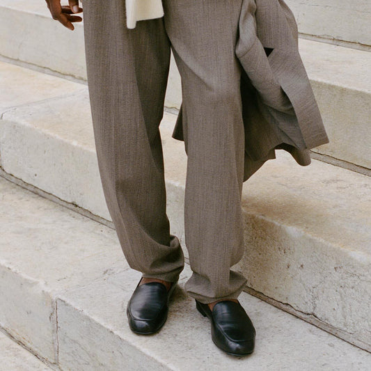 A person in loose brown pants and black shoes stands on stone steps, holding a matching brown jacket. Their upper body and face are not shown in the image. The sleek black shoes are The James Loafer - Black Nappa by Margaux, an everyday loafer crafted with sacchetto construction for exceptional comfort.