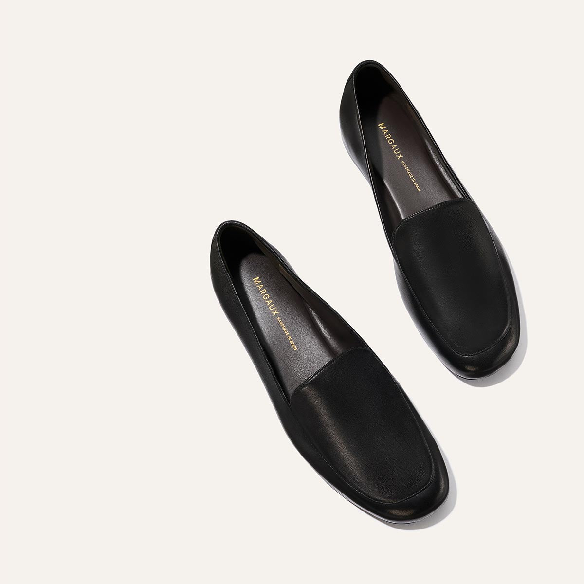 The James Loafer - Black Nappa by Margaux is shown on a light beige background, angled to display the insoles with "Margaux" printed in gold inside each shoe.