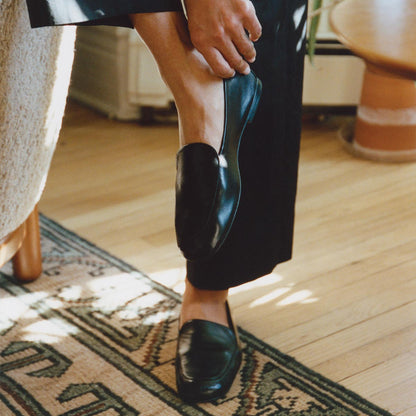 A person wearing Margaux's The James Loafer in Black Nappa stands on a wooden floor by a patterned rug, lifting one pant leg to reveal the shoe. Warm sunlight fills the room, highlighting its slipper-like comfort.