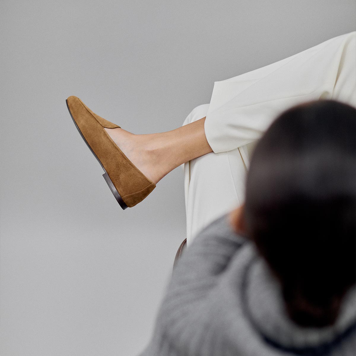 A person sits with one leg crossed, viewed from behind against a neutral background, wearing white pants and Margaux’s The James Loafer in Cedar Suede.