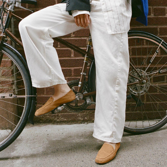 Wearing crisp white pants and Margaux’s The James Loafer in Cedar Suede, a person stands next to a brown bicycle with one foot on the pedal, set against a brick wall.