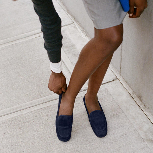 Standing on a sidewalk with the wall in the background, a person wearing a grey skirt and dark sweater bends down to adjust Margaux’s The James Loafer in Indigo Suede—handmade with sacchetto construction—on their foot.