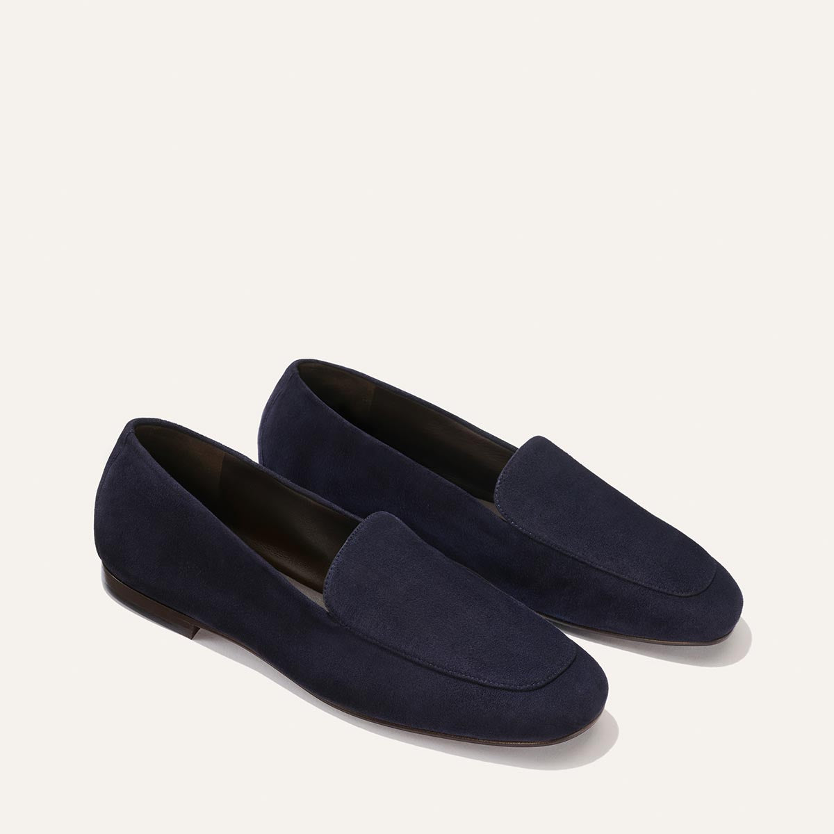 The James Loafer in Indigo Suede by Margaux features a refined, slip-on silhouette and sacchetto construction for a tailored fit, elegantly presented against a light beige background.