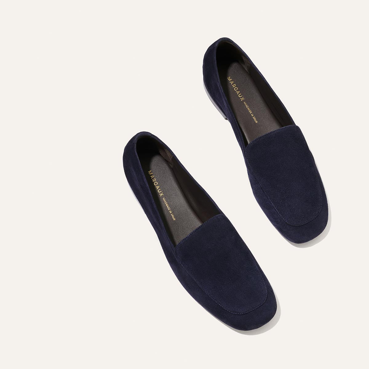 A pair of The James Loafer - Indigo Suede by Margaux, featuring a minimalist design and tailored fit, are displayed on a light beige background, positioned slightly apart.