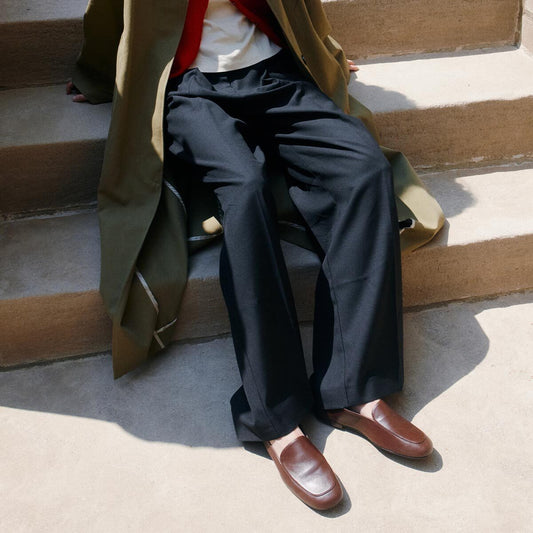 Seated on outdoor steps, the individual—face turned away from view—wears a long olive coat, red sweater layered over a white top, wide black pants, and Margaux’s The James Loafer in Mahogany Nappa, renowned for their handcrafted sacchetto construction.