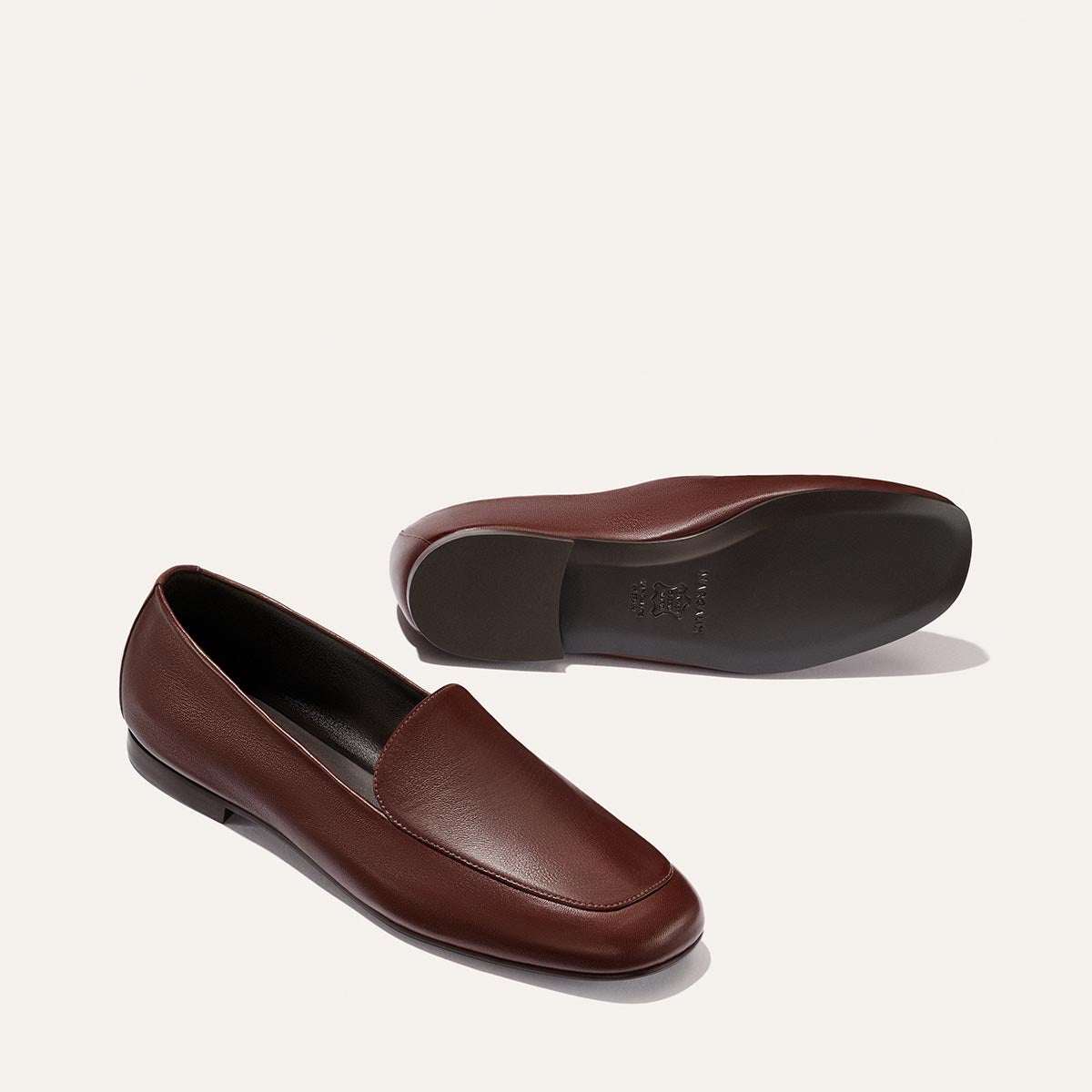 A pair of Margaux's The James Loafer in Mahogany Nappa, designed in the sacchetto loafer style for slipper-like comfort, shown with one shoe standing upright and the other lying on its side to reveal the sole, set against a plain light background.