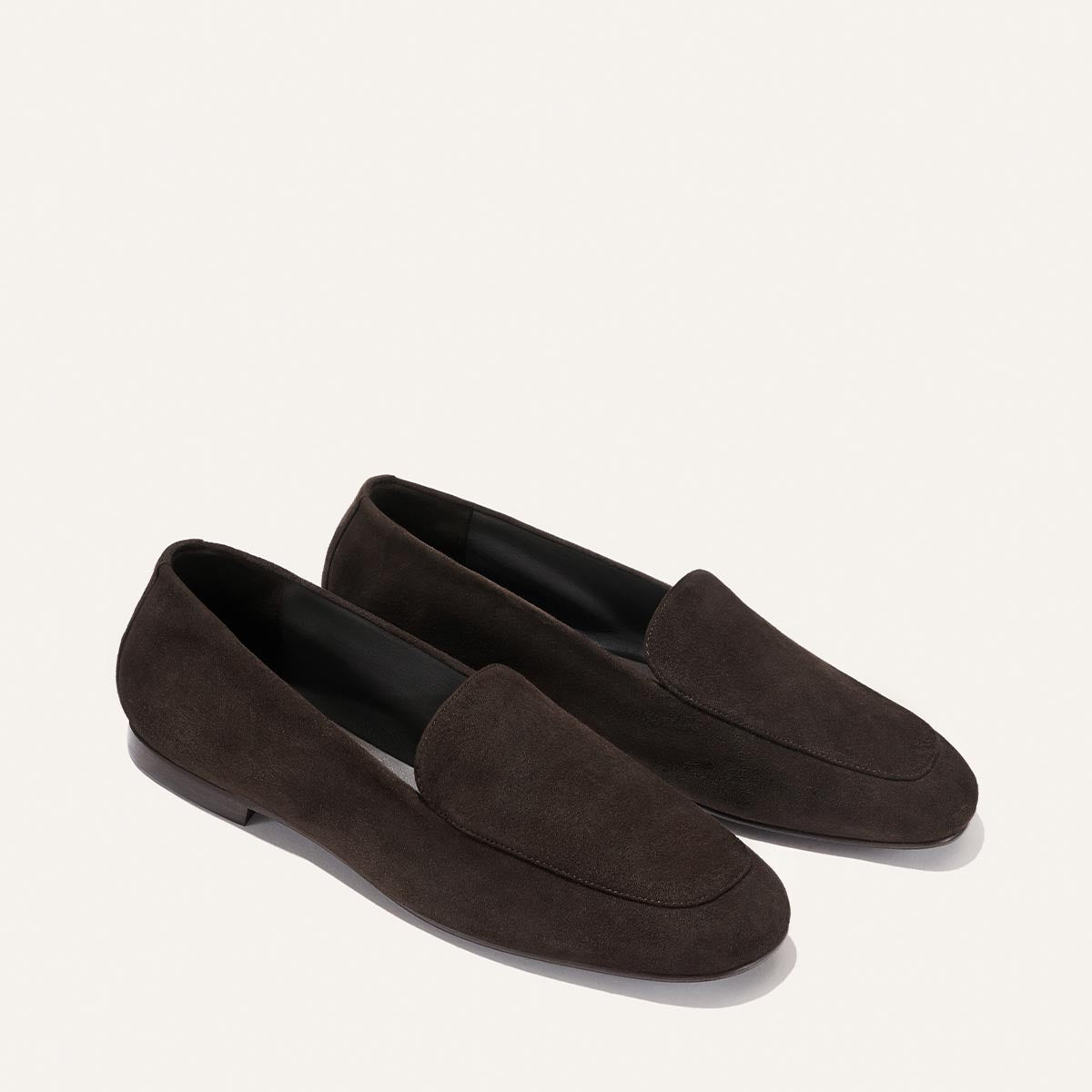 The James Loafer - Molasses Suede by Margaux features a simple, classic design with flat soles and rounded toes, crafted in dark brown suede. Offering slipper-like comfort, these loafers are elegantly showcased on a light background.