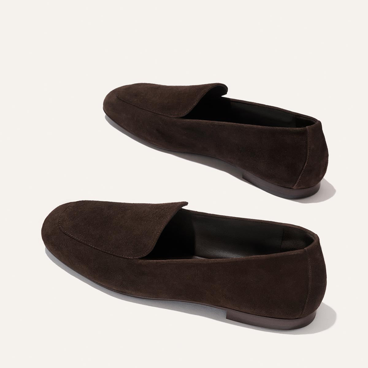 The James Loafer - Molasses Suede by Margaux is a pair of dark brown suede loafers featuring a sleek, slip-on design and flat soles, meticulously crafted with sacchetto construction for a tailored fit, beautifully displayed on a light background.