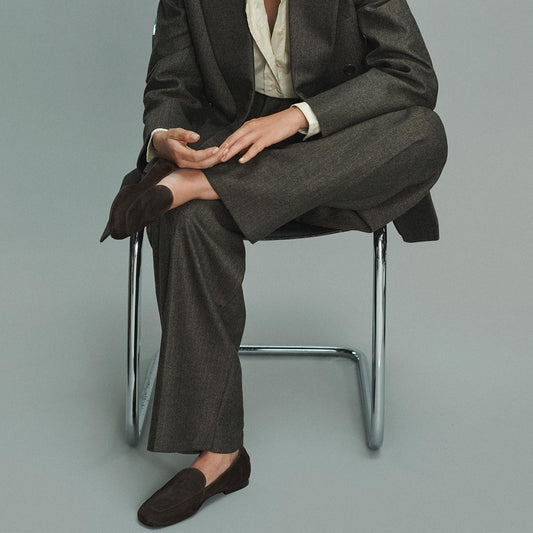 A person in a dark gray suit sits on a metal chair with legs crossed, wearing the Margaux James Loafer in Molasses Suede and a light-colored shirt. The person's face is not visible.