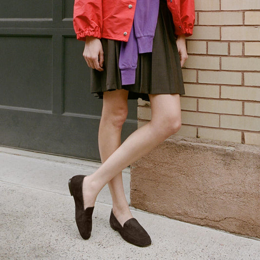 A person in a red jacket and black skirt stands with legs crossed, wearing Margaux’s The James Loafer in Molasses Suede, next to a brick wall and green garage door.