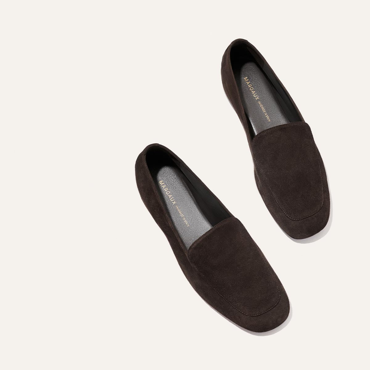 A pair of Margaux's The James Loafer in Molasses Suede, featuring a minimalist design and slipper-like comfort, is displayed diagonally on a light beige background, with one handmade loafer positioned slightly above the other.
