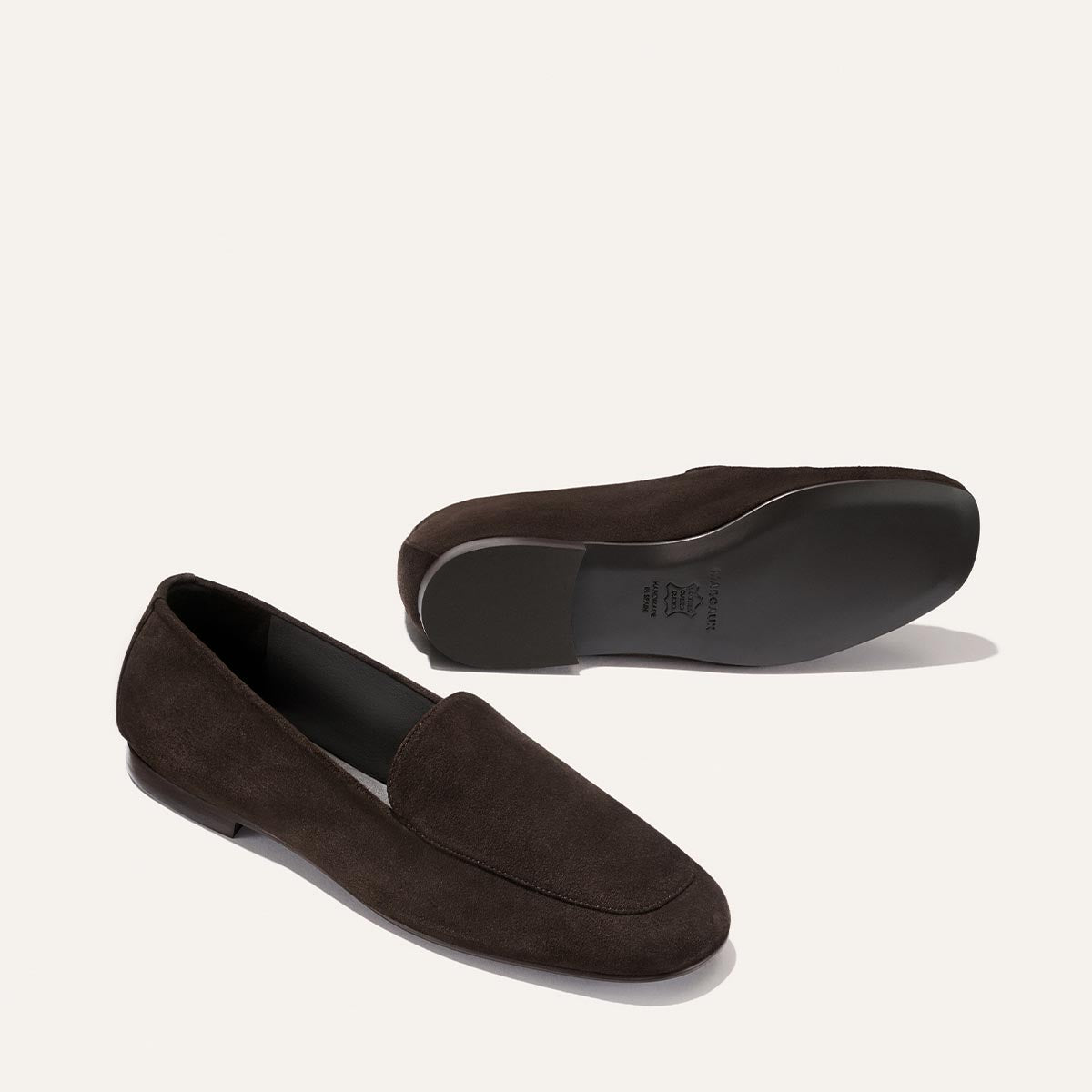 A pair of Margaux’s The James Loafer in Molasses Suede is displayed, with one shoe upright and the other positioned to showcase the sole. Renowned for their slipper-like comfort and expert handmade craftsmanship, these loafers are set against a plain white background.