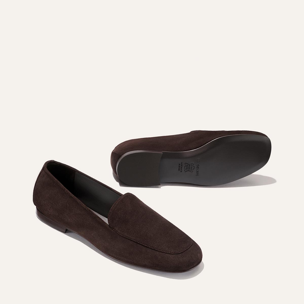 A pair of The James Loafer in Molasses Suede by Margaux—expertly handcrafted with sacchetto construction for a bespoke fit—are displayed: one shoe standing tall and the other resting on its side to highlight the smooth, sculpted sole of this artisanal loafer.