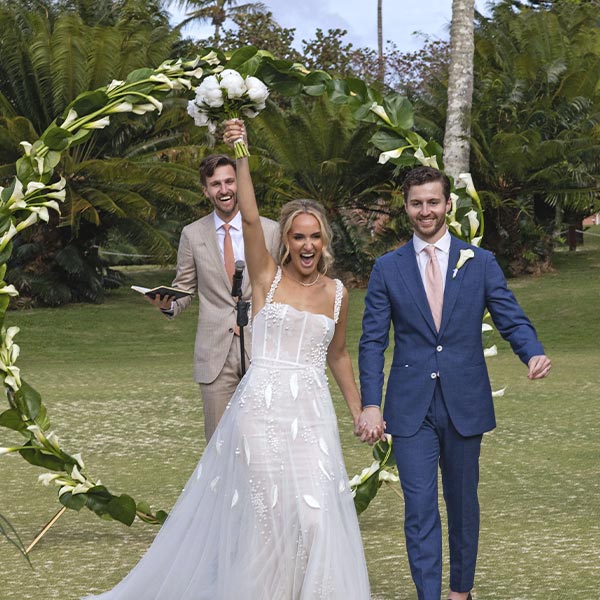 A joyful bride in a white dress holds a bouquet triumphantly as she walks hand in hand with the groom, both smiling, in front of a circular floral arch outdoors, with an officiant standing behind them.