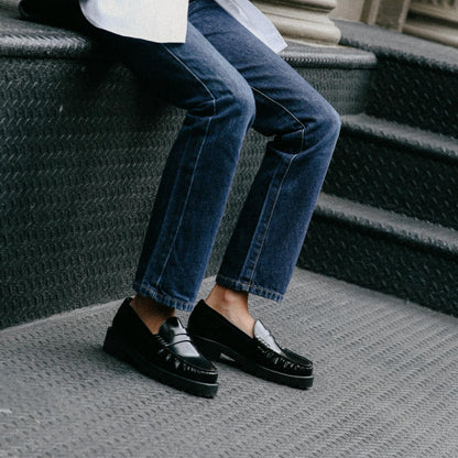 A person in blue jeans and shiny Margaux Louisa Loafer First Edition - Black Calf sits on outdoor metal steps, legs and feet visible to showcase the elegant leather-lined lug-sole loafer design.