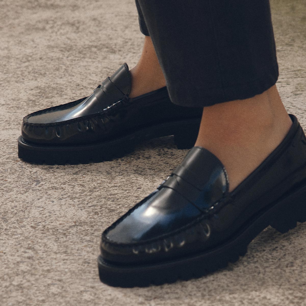 A close-up of a person wearing Margauxs The Louisa Loafer in Black Calf and dark pants, standing on a light textured surface, highlights this stylish shoe’s comfortable fit.