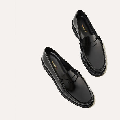 The Louisa Loafer First Edition - Black Calf by Margaux, featuring shiny black uppers and thick, textured lug soles, is displayed on a light background. Leather insoles bear the Margaux brand name in gold lettering.