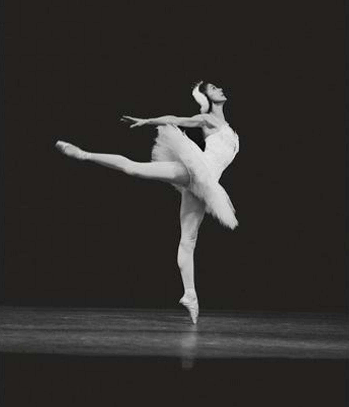 A ballerina in a tutu and headpiece gracefully balances on one toe with one leg extended behind her, performing on stage against a dark background.