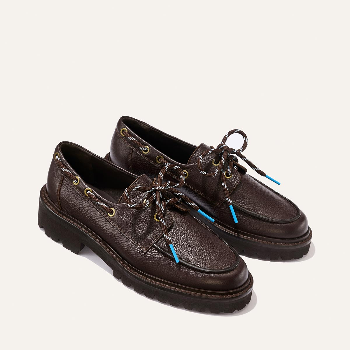 The Marine Loafer - Espresso Tumbled Calf by Margaux is a pair of dark brown leather boat shoes with a textured tumbled finish, thick black soles, gold eyelets, and multicolored laces with blue aglets, shown against a light background.