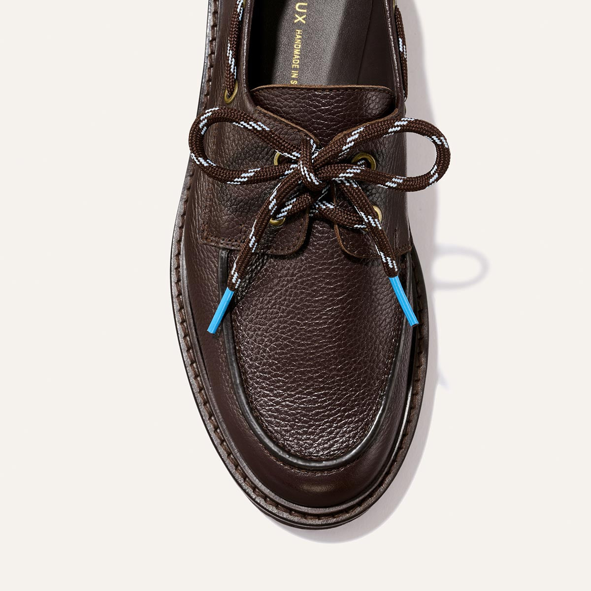A top view of The Marine Loafer – Espresso Tumbled Calf by Margaux showcases its brown textured leather, brown-and-white laces tied in a bow with blue lace tips, set against a light background.
