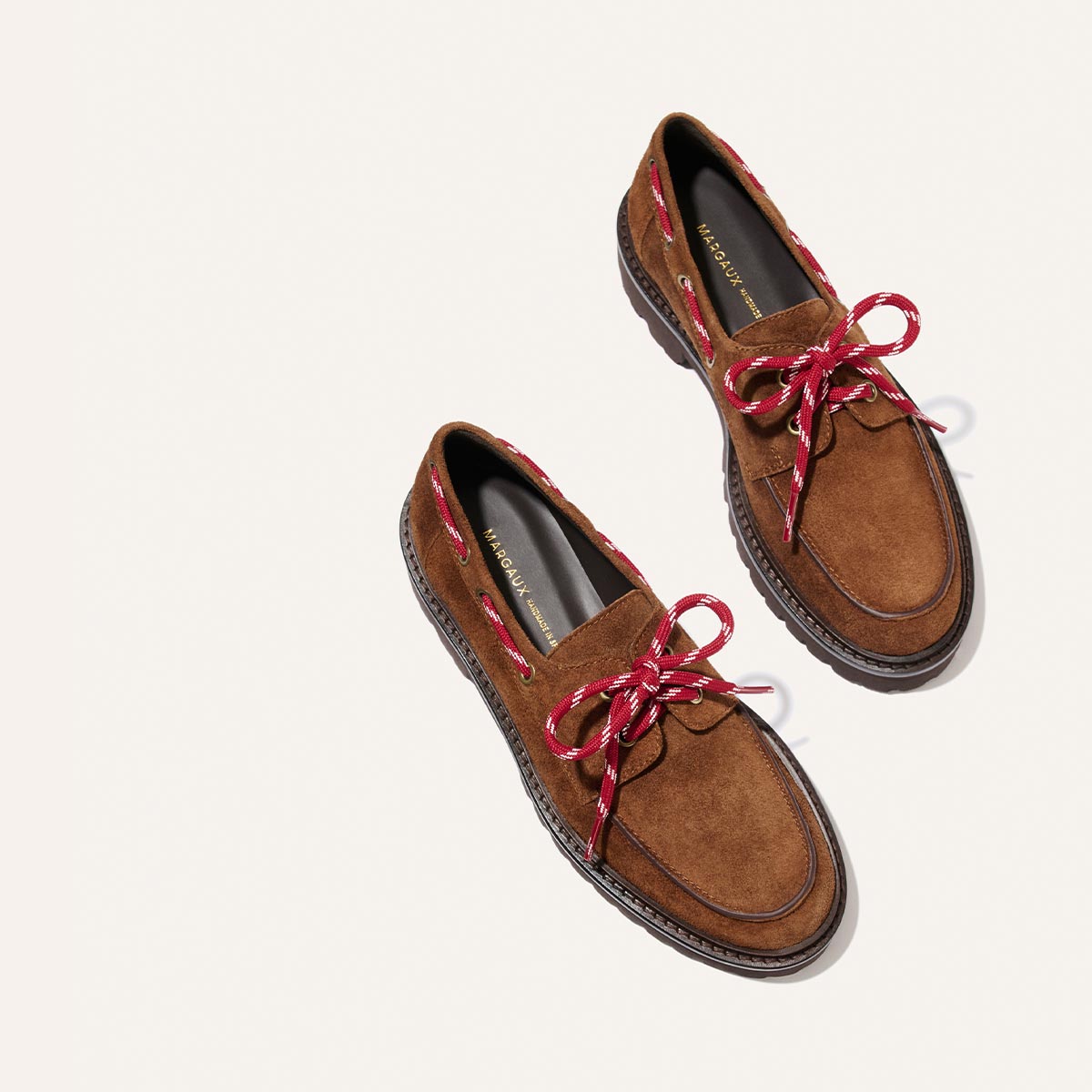 The Marine Loafer – Saddle Suede by Margaux, featuring brown suede uppers, dark brown soles, and red and white patterned laces, is displayed on a light background with prominent stitching around the edges.