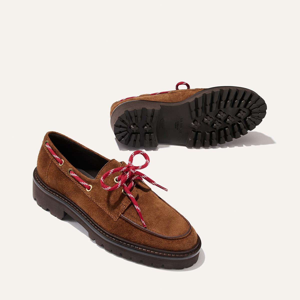 A pair of The Marine Loafer - Saddle Suede by Margaux, featuring brown suede uppers, chunky black soles, and red-and-white striped laces—one shoe displayed upright and the other showcasing the unique tread pattern on the sole.