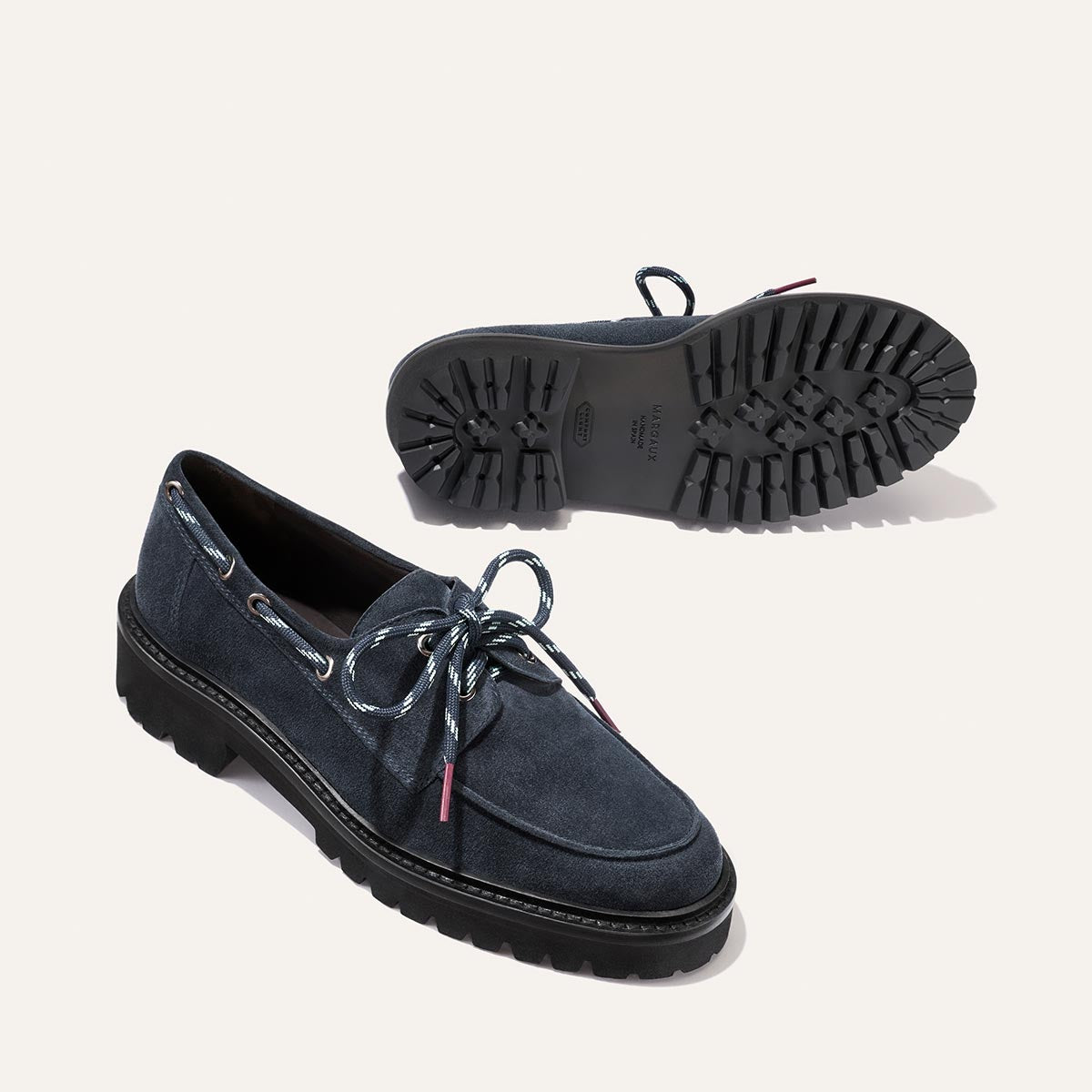 A pair of Margaux’s The Marine Loafer in Vintage Navy Suede, featuring thick black rubber soles, blue and white laces threaded through silver eyelets. One loafer stands upright while the other rests on its side, highlighting the unique tread pattern.