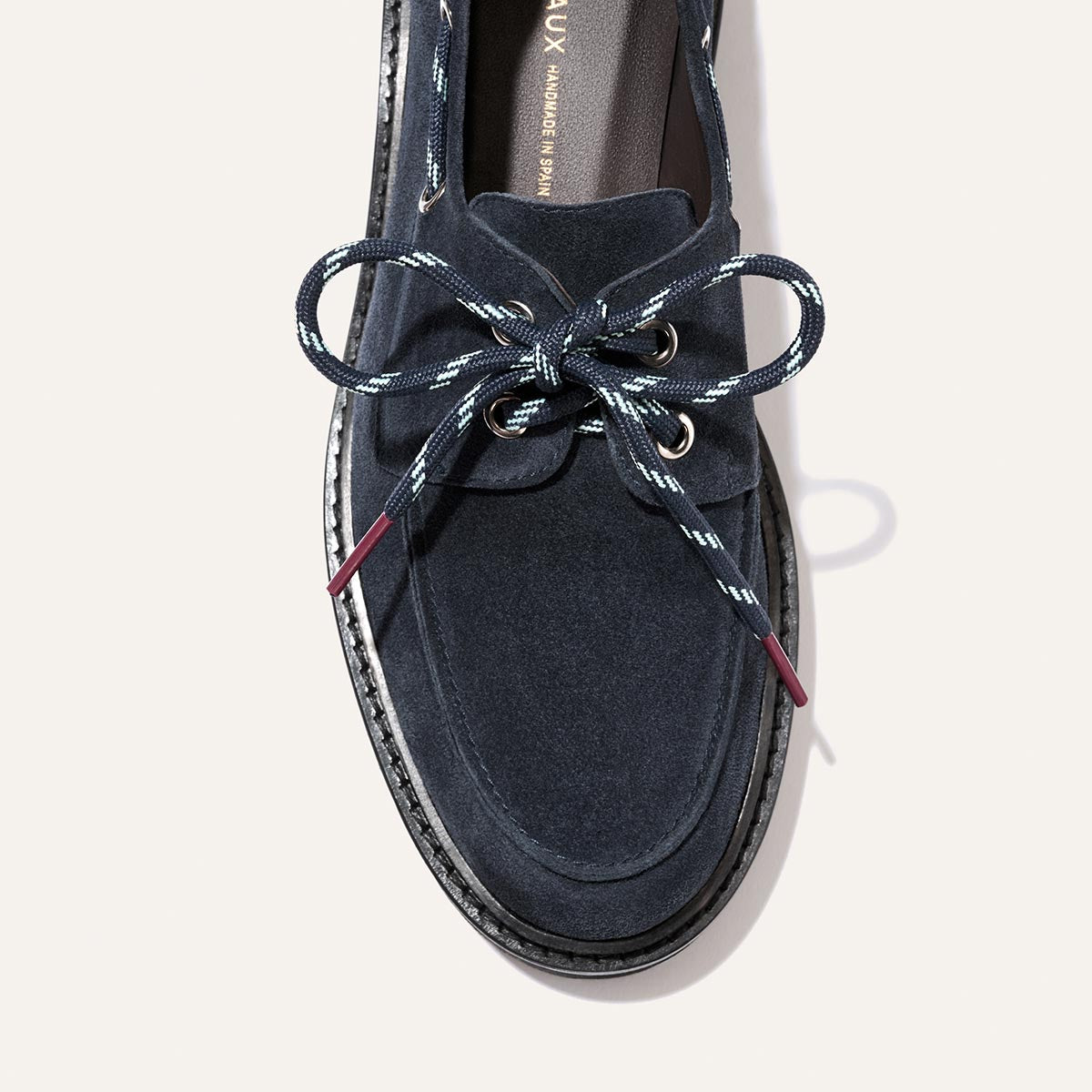 Introducing The Marine Loafer by Margaux in Vintage Navy Suede—a sophisticated navy blue suede boat shoe featuring crisp white-stitched accents, metal eyelets, and dark blue laces finished in a neat bow. This elegant loafer rests on a dark sole and stands out beautifully against a light background.