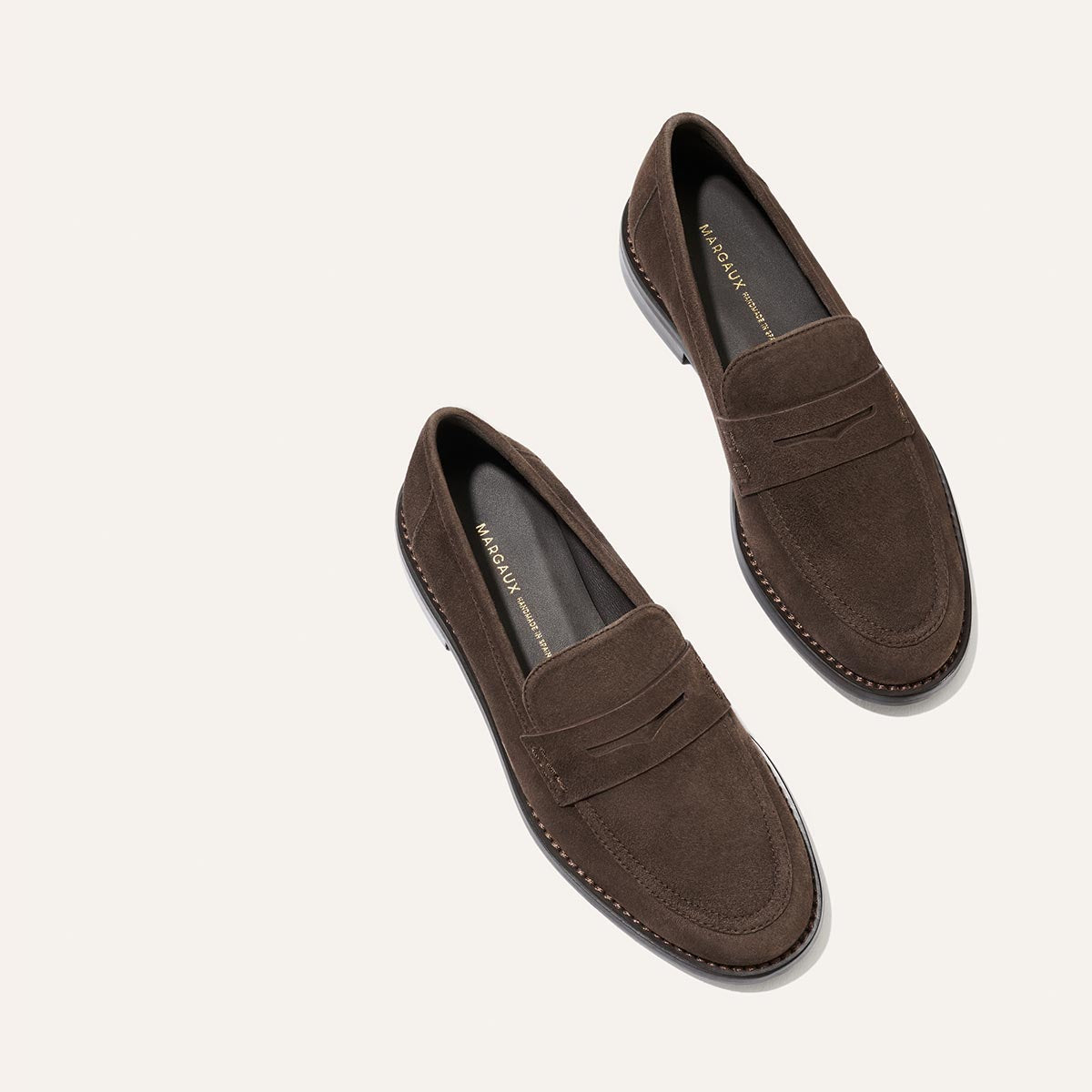 The Marlowe Loafer by Margaux in Umber Suede features refined stitching details and contrasting black soles, elegantly displayed at an angle on a minimalist light background—a timeless essential for the modern loafer enthusiast.