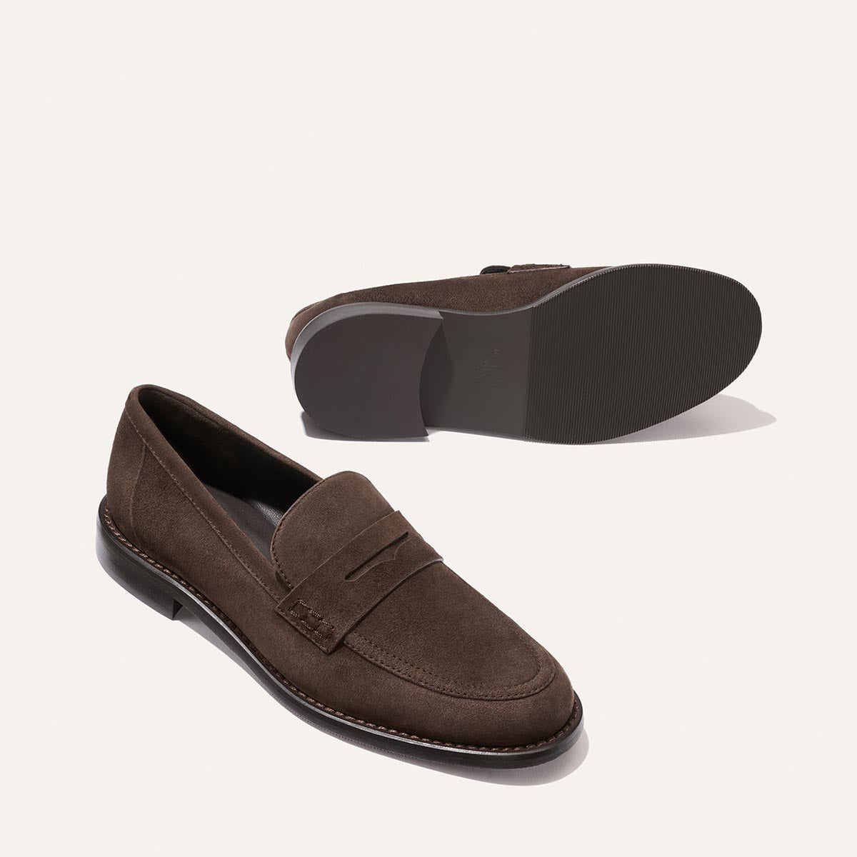 A pair of The Marlowe Loafer in Umber Suede by Margaux—a timeless closet staple—are shown against a plain light background, with one shoe upright to reveal the top and side, and the other lying on its side to display the sole.